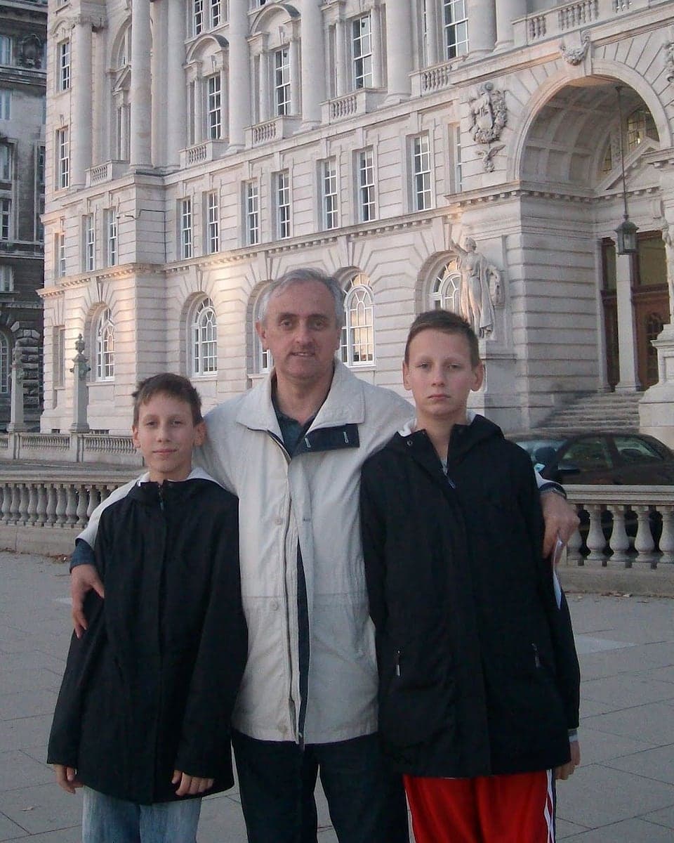 Andrija T. Sarić u svetloj jakni stoji između svoja dva sina u crnim jaknama, ispred kamene zgrade sa stubovima i lukovima u Liverpulu.